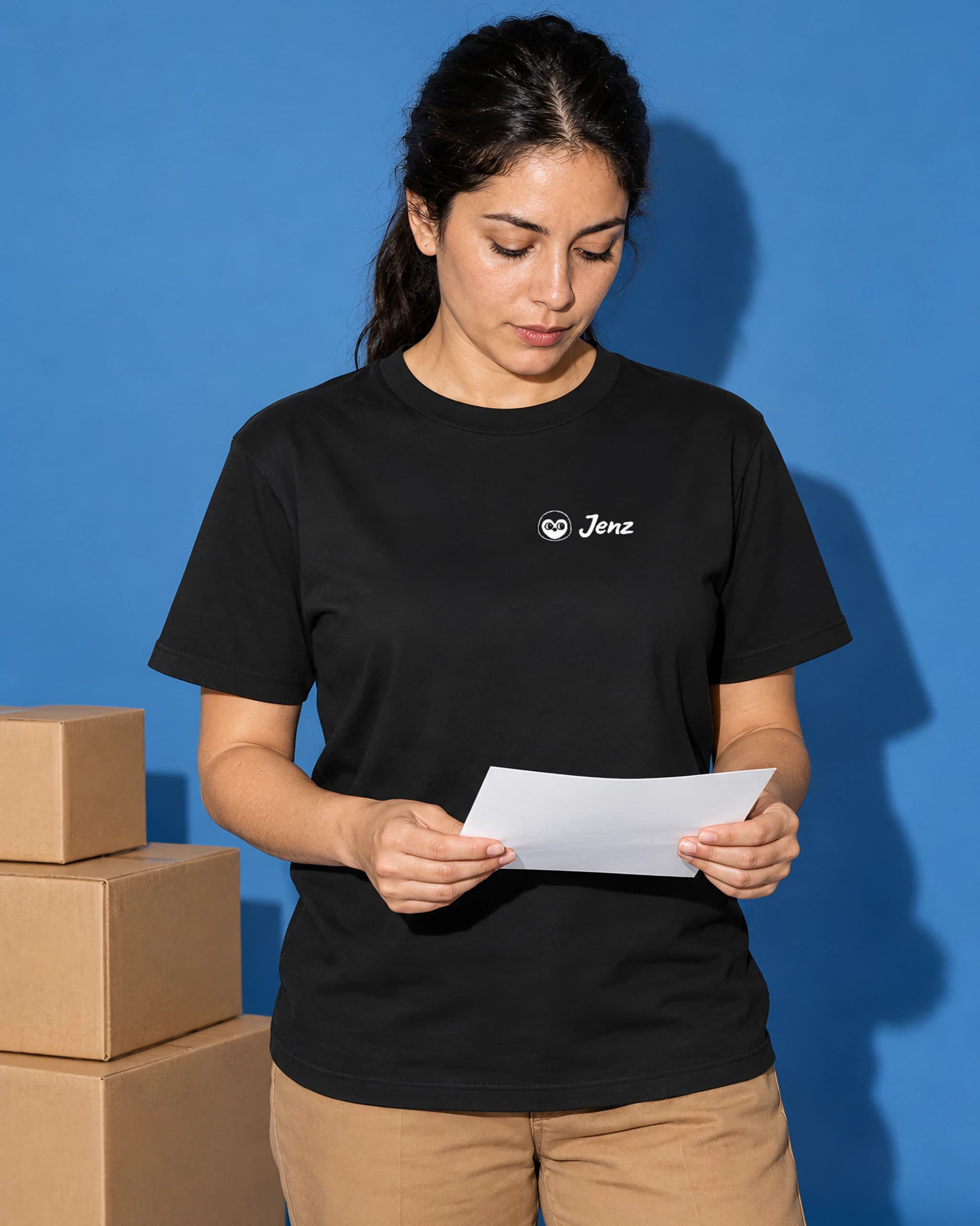 Woman in a black T-shirt with a small white Jenz logo, reading a note beside stacked boxes
